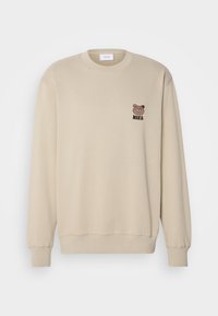 Felpa beige con scollo rotondo, polsini e orlo a costine. Presenta un piccolo ricamo di un orsetto bruno e il testo "MAKIA" sul petto.