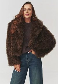 Brązowa kurtka z futra syntetycznego o shaggy teksturze, z szerokim kołnierzem i skróconymi rękawami, noszona na ciemnym swetrze i wysokich dżinsach.