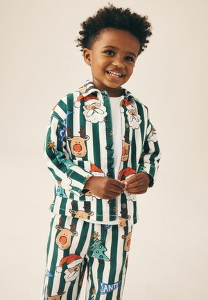 Niño pequeño sonriente vestido con un conjunto de rayas verdes y blancas con estampados de Papá Noel, renos y árbol de Navidad.