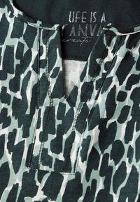 Kortärmad plagg i grönt och vitt djurtryck. Har en u-ringning och synliga sömmar. Materialet verkar mjukt och texturerat.