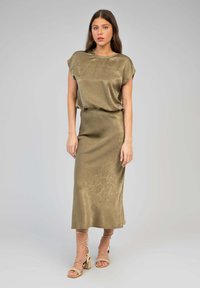 Robe midi en satin vert olive avec des manches courtes, coupe ample et texture lisse. Associée à des sandales à talons carrés beiges à lanières.