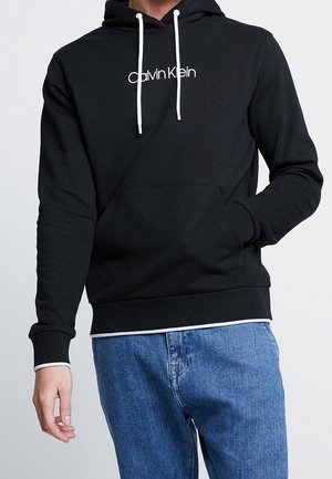 Czarna bluza z kapturem Calvin Klein z białymi sznureczkami noszona z niebieskimi dżinsami, ręka w przedniej kieszeni, styl casual.