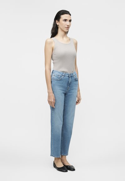 Γυναίκα που στέκεται αντικρίζοντας μπροστά, φορώντας μπεζ αμάνικο τοπ, ανοιχτό μπλε ψηλόμεσα τζιν και μαύρα ίσια παπούτσια μπροστά σε απλό φόντο.