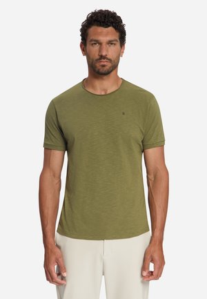 Man met krullend haar en baard, die een olijfgroen T-shirt met korte mouwen en lichtbeige broek draagt, staand tegen een effen achtergrond.