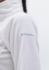 Gros plan sur la manche d'une veste en polaire blanche avec un logo Columbia brodé visible près de l'épaule.