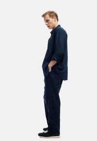 Marineblaues Hemd mit lockerer Passform und langen Ärmeln, kombiniert mit passenden marineblauen Hosen. Schwarze Sneakers mit weißen Schnürsenkeln sorgen für einen Kontrast.