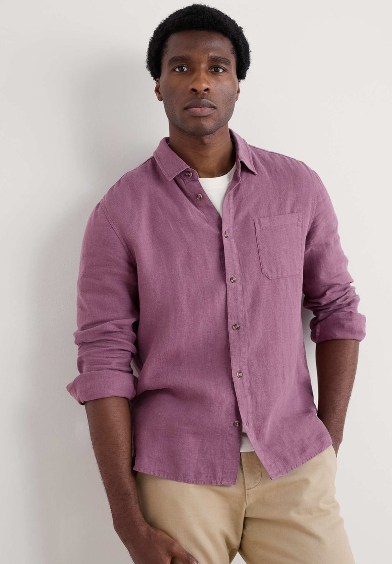 Camisa de lino en suave color malva con cuello abotonado, bolsillo en el pecho, mangas arremangadas y botones de madera. Se lleva sobre una camisa blanca.