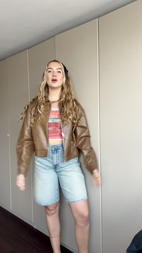 Chaqueta bomber corta de cuero marrón sobre un top crop de rayas multicolores, combinada con pantalones cortos de mezclilla azul claro, de pie contra una pared neutra.