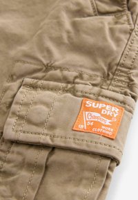 Bolsa de tela marrón con etiqueta naranja "Superdry", que presenta detalles de costura y un diseño de vestuario de trabajo. Superficie compacta y texturada.
