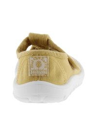Zapatilla de lona amarilla con suela blanca y correa trasera. Presenta un parche con el logo en el talón y un diseño texturizado para agarre y soporte.