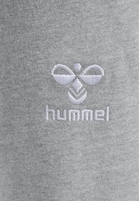 Białe haftowane logo pszczółki Hummel oraz nazwa marki na jasnoszarym tle z tkaniny.