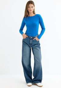 Top de manga larga azul brillante combinado con jeans de mezclilla de pierna ancha y cintura alta. Textura de tela suave y cierre de botones visible en la cintura.