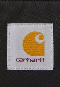 Biały materiałowy metka z pomarańczowym logo Carhartt i ciemnobrązowym tekstem wszytym na czarnym teksturowanym tle.