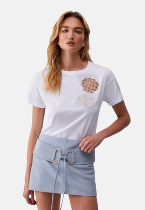 Donna che indossa una camicia bianca a maniche corte con ricamo a conchiglia beige e una minigonna azzurra con cintura, in piedi davanti a uno sfondo uniforme.