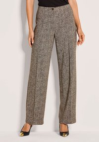 Vrouw in brede bruine broeken met een visgraatpatroon en knoopsluiting, gecombineerd met zwarte puntige hakken met gouden tips.