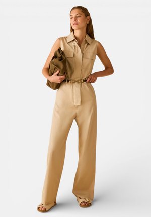 Donna con tuta beige senza maniche e cintura, che tiene una borsa marrone oversize, in piedi con una mano sui fianchi, guardando di lato.