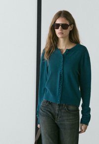 Cardigan turchese realizzato in morbido tessuto a maglia, con scollo rotondo, chiusura frontale con bottoni e polsini a coste, indossato con jeans in denim scuro.