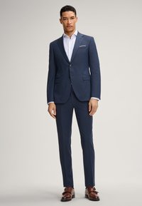 Abito tailored blu navy con vestibilità slim, caratterizzato da rever a scollatura, chiusura a un bottone e texture liscia. Scarpe in pelle marrone completano il look.