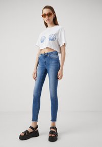 Modré džínové skinny džíny s vybledlým vzhledem, v kombinaci s bílým zkráceným tričkem se dvěma modrými mušlemi. Černé platformové sandály.