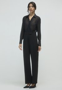 Femme debout portant un blouse en dentelle noire à manches longues, un pantalon noir à taille haute et jambes larges, des talons aiguilles noirs et des boucles d'oreilles pendantes.