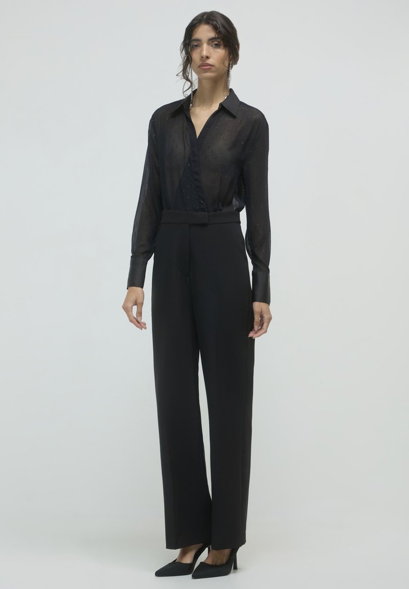 Femme debout portant un blouse en dentelle noire à manches longues, un pantalon noir à taille haute et jambes larges, des talons aiguilles noirs et des boucles d'oreilles pendantes.