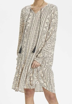 Robe beige à manches longues avec des motifs géométriques noirs, un décolleté en V, des détails à franges et un ourlet à volants, fabriquée en tissu léger.