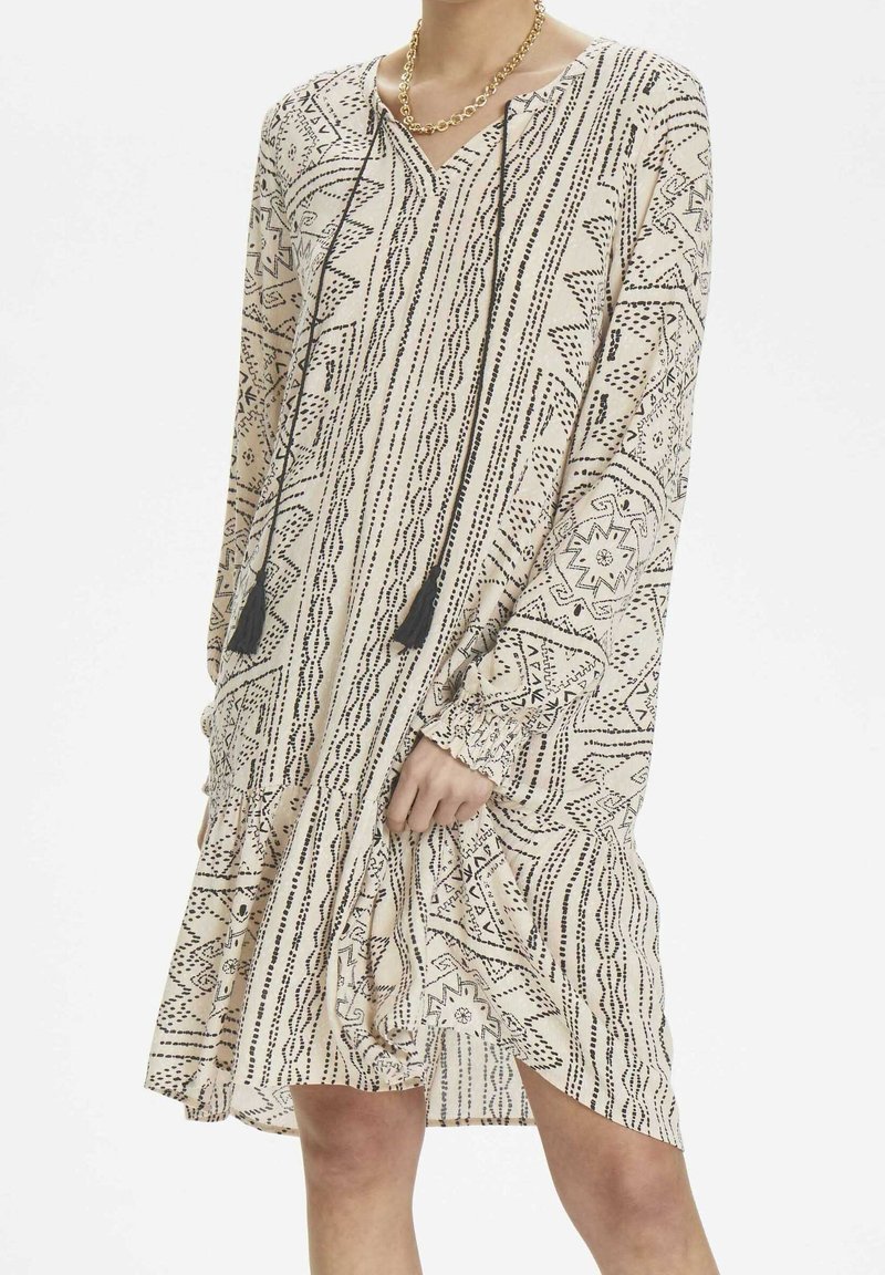 Långärmad, beige klänning med svarta geometriska mönster, v-ringning, fransdetaljer och en volangkjol, tillverkad av lättviktsmaterial.