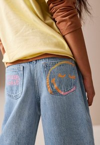 Lys gul skjorte lagdelt over brune ærmer, kombineret med blå denimjeans med pink og orange smilende ansigt og "Smiling World"-design på lommerne.