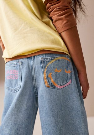 Heledat kollast särki kantakse pruunide varrukate kohal, sellega kaasnevad sinised teksapüksid, mille taskutel on roosade ja oranžide naeratavate nägude ning "Smiling World" kujundused.