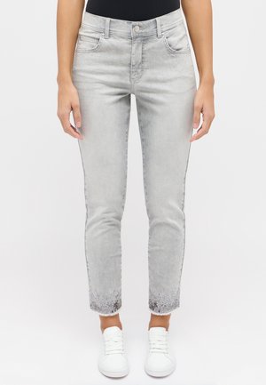 Hellgraue Denim-Jeans mit taillierter Passform, fransigem Saum und verzierten Enden mit silbernen Akzenten an den Bündchen. Getragen mit weißen Sneakers.