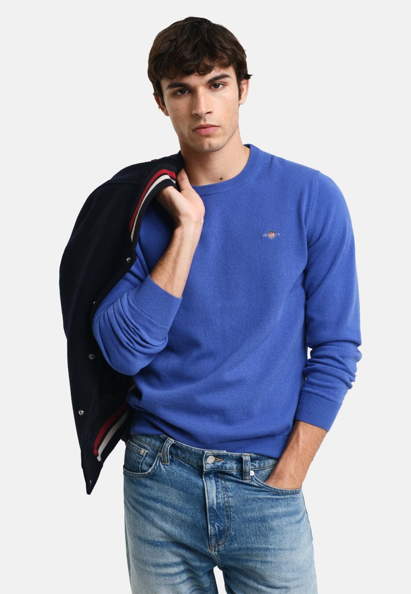 Maglione blu a maglia con scollo rotondo e polsini a coste, indossato con denim blu casual. Un piccolo logo arricchisce la zona del petto.