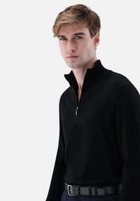 Schwarzer Reißverschluss-Pullover aus weichem Stoff, mit hohem Kragen und figurbetontem Design. Wird über dunklen Hosen getragen und durch einen schwarzen Gürtel ergänzt.