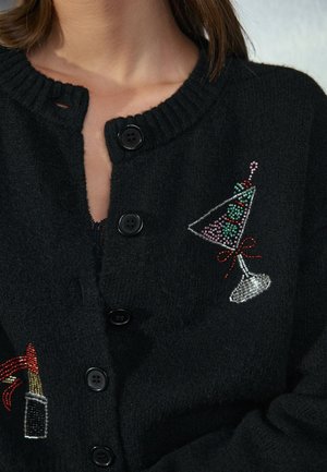 Must kardigan mustade nööpidega, mida kannab inimene, mille rinnal on tikitud kokteiliklaas värviliste helmestega ja vöökohal huulepulga kujundus.