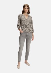 Kobieta w beżowej i czarnej bluzce w zebrę, jasnoszarych skinny jeansach i beżowych butach typu slip-on, stojąca z jedną ręką w kieszeni.