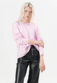 Een model met lang, steil blond haar draagt een roze gebreide trui en zwarte leren broek, en poseert zelfverzekerd tegen een witte achtergrond.