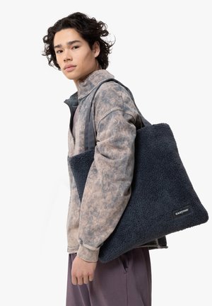 Tote bag en polaire bleue avec une surface texturée. Tissu doux, forme rectangulaire et poignées robustes. Présente une étiquette noire avec le texte "EASTPAK".