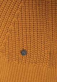 Kudottu sinappinvärinen pipo, jossa on teksturoitu kuvio ja ribbireunus. Edessä metallinen logo-etiketti yksityiskohtana.