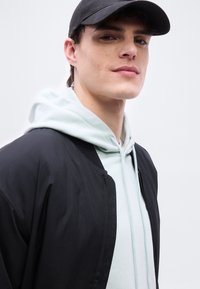 Νέος άνδρας που φοράει μαύρο καπέλο, ανοιχτό γκρι φούτερ με κουκούλα και μαύρο μπουφάν, κοιτάζει με αυτοπεποίθηση την κάμερα σε απλό φωτεινό φόντο.