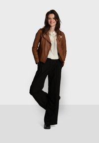 Veste de moto en cuir marron sur une blouse blanche, associée à un pantalon large noir et des chaussures noires, modèle debout avec une jambe croisée.