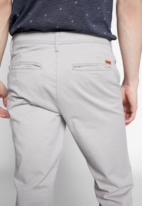 Pantalon gris clair à texture lisse, doté de deux poches arrière et d'une petite étiquette logo sur la poche droite. Design décontracté, coupe ajustée.