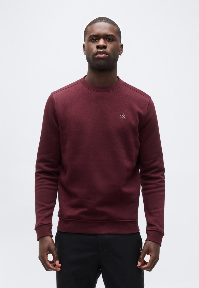 Νέος άνδρας όρθιος, φοράει μακρυμάνικο πουλόβερ Calvin Klein σε μπορντό χρώμα και μαύρο παντελόνι, μπροστά σε απλό φωτεινό φόντο.