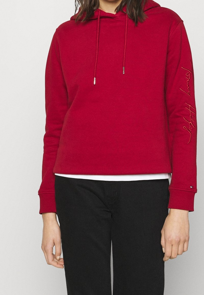 Personne portant un sweat à capuche rouge avec cordons, pantalon noir et un t-shirt blanc visible sous l'ourlet.