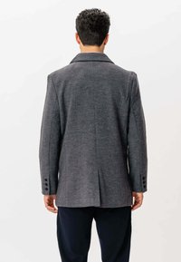 Mand iført et mørkegråt skræddersyet blazer med lukkede manchetter, set fra ryggen mod en plain lys baggrund.