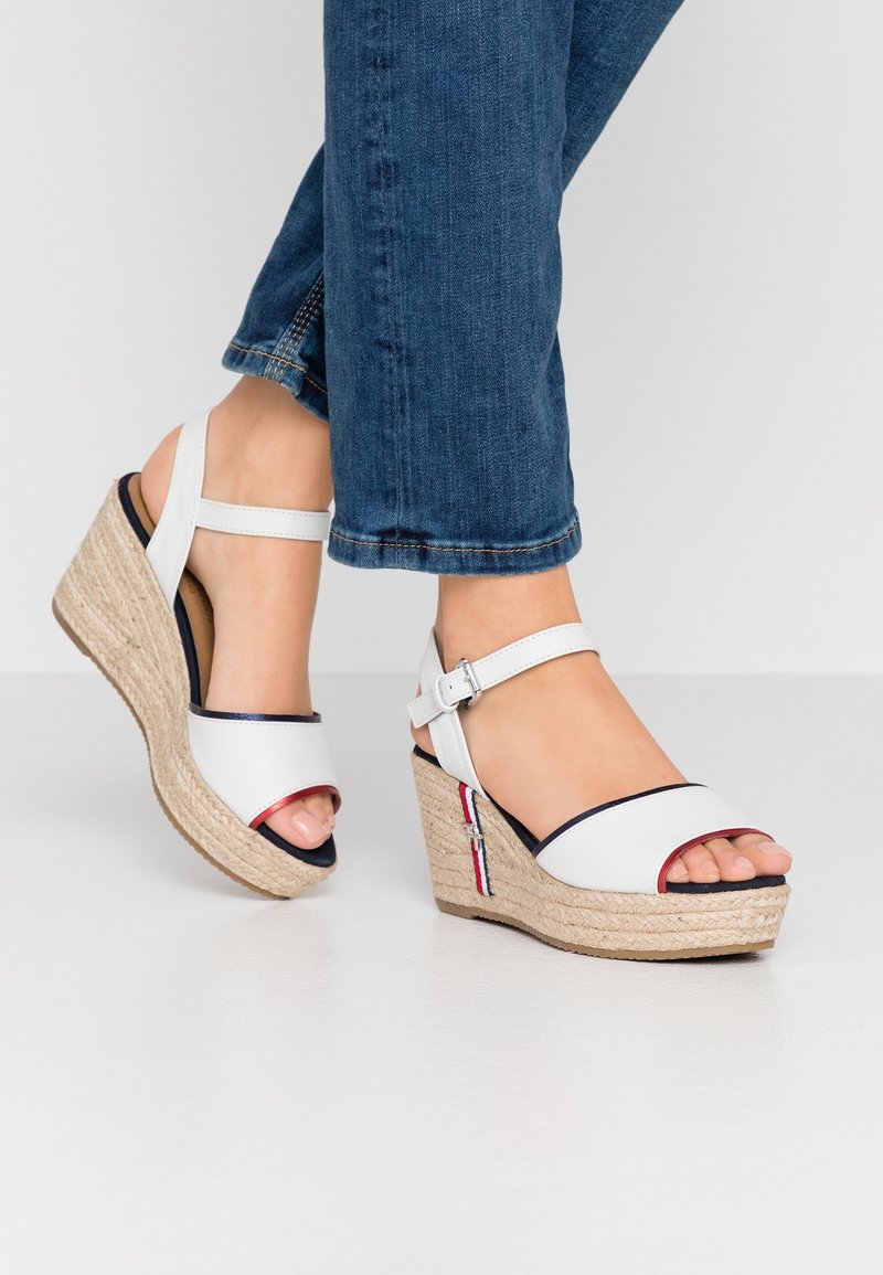 Vita klacksandaler med en vävd juteplattform, ankelrem, öppen tå och röd/blå accentdetaljer. Bärs med mörk denimjeans.