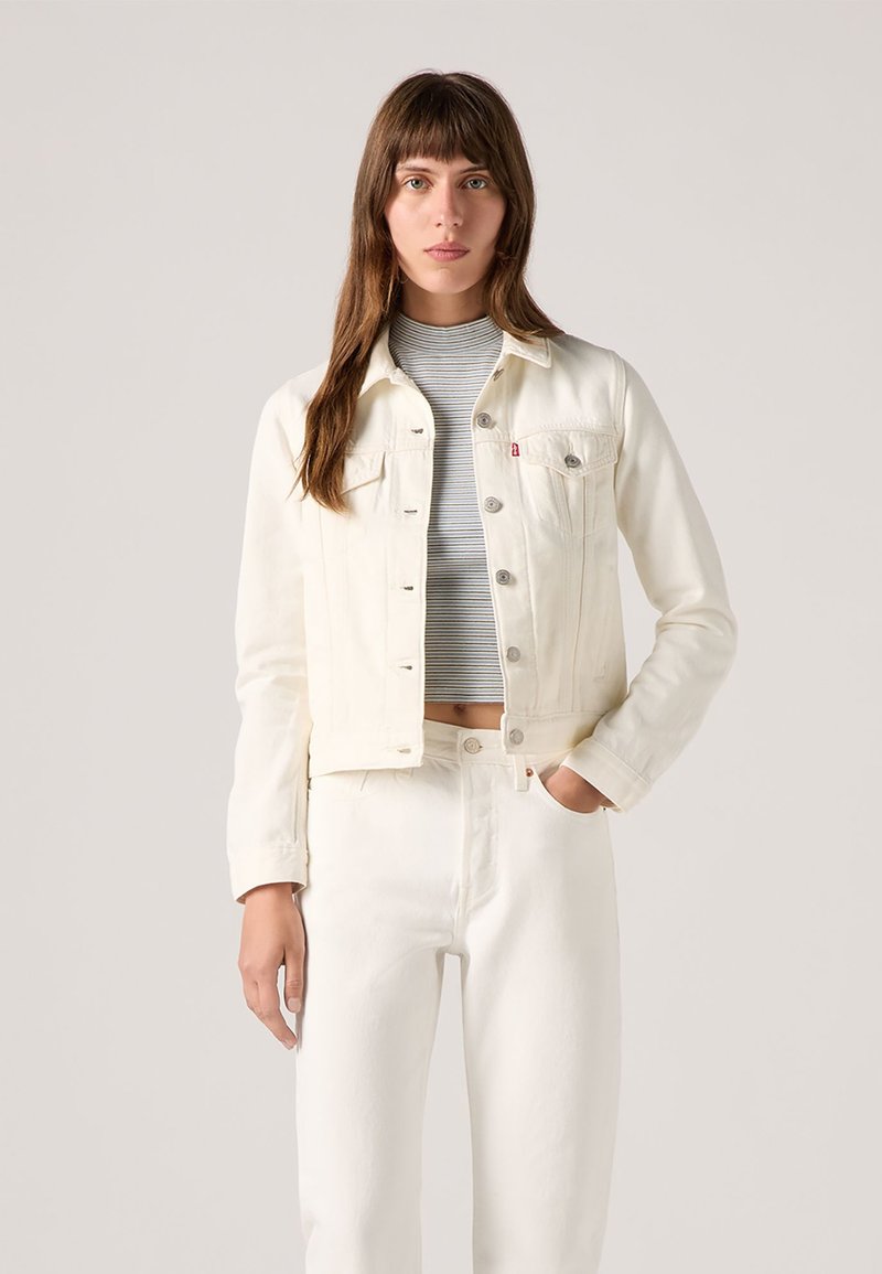 Chaqueta de mezclilla blanca con botones plateados, bolsillos en el pecho y un diseño ajustado, combinada con pantalones blancos a juego y una blusa a rayas.