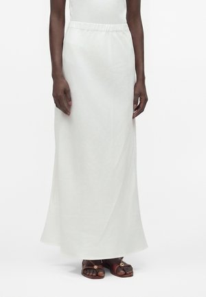 Personne portant une jupe blanche longue jusqu'aux chevilles avec une taille élastique et des sandales marron à bout ouvert avec de petites ornements dorés.
