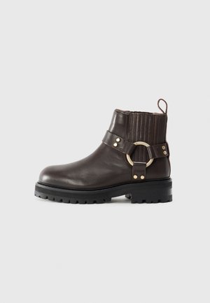 Stivale alla caviglia in pelle marrone con pannelli laterali elasticizzati, accentato da un anello dorato e una suola nera spessa. Presenta dettagli brogue sulla parte superiore.