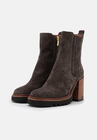 Botas de tobillo de ante gris oscuro con un tacón grueso, cremallera lateral y detalles acanalados verticales. Presentan una suela texturizada y un ribete marrón en contraste.