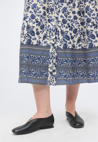 Κάτω άκρα με μαύρα χαμηλά παπούτσια slip-on και μια μπεζ φούστα με μπλε ανθισμένα και ριγέ μοτίβα, που στέκονται σε λευκό πάτωμα.