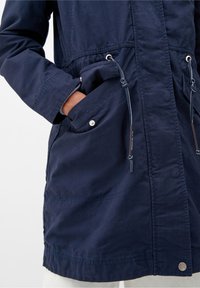 Chaqueta azul marino con cierre de cremallera en el frente, cintura con cordón, grandes bolsillos frontales y botones a presión plateados. Fabricada con una tela suave con una textura sutil.